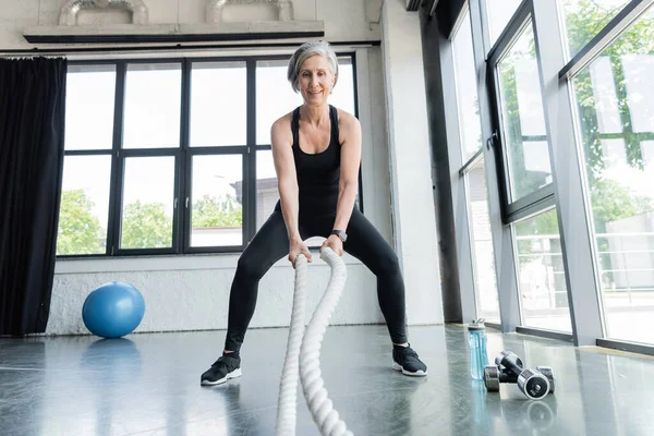 Pleine longueur de femme âgée positive travaillant avec des cordes de combat près des haltères dans la salle de gym — Photo de stock