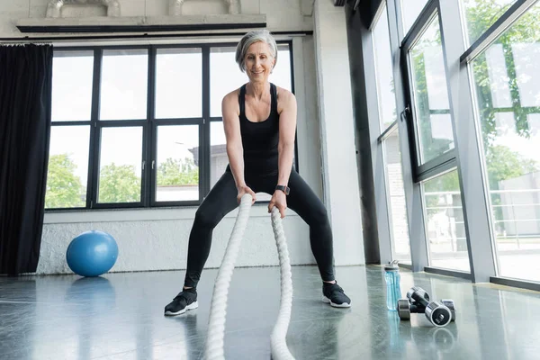 Longitud completa de la mujer mayor en forma haciendo ejercicio con cuerdas de batalla cerca de pesas en el gimnasio - foto de stock