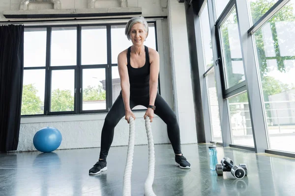 Full length of happy senior woman training with battle ropes near dumbbells in gym — Stock Photo