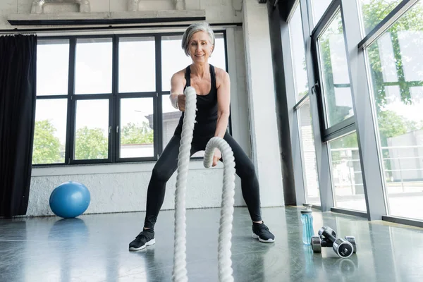 Longitud completa de la mujer mayor alegre entrenamiento con cuerdas de batalla cerca de pesas en el gimnasio - foto de stock