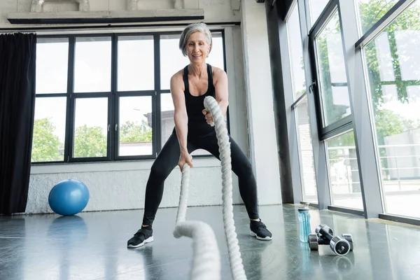 Longitud completa de entrenamiento positivo de la mujer mayor con cuerdas de batalla cerca de pesas en el gimnasio - foto de stock