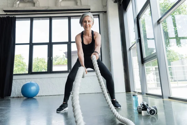 Full length of positive senior woman working out with battle ropes in gym — Stock Photo