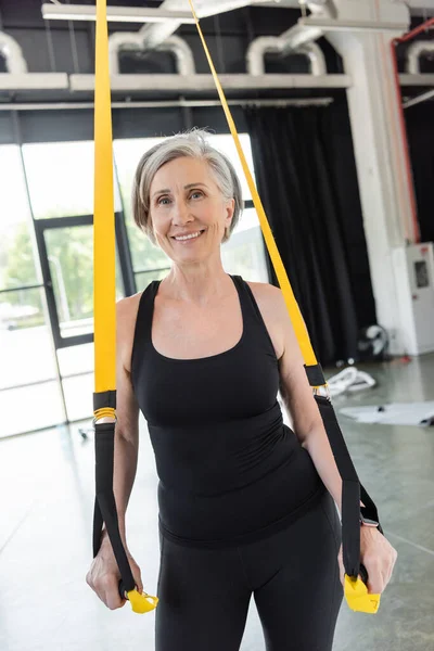 Heureuse femme âgée en noir sportswear exercice avec des bretelles de suspension dans la salle de gym — Photo de stock