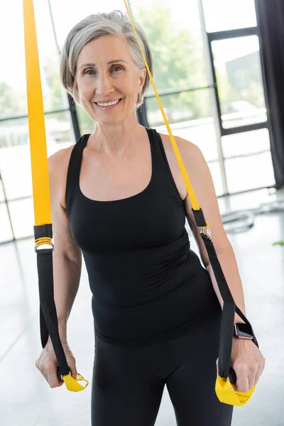 Gaie femme âgée en noir vêtements de sport exercice avec des bretelles de suspension dans la salle de gym — Photo de stock