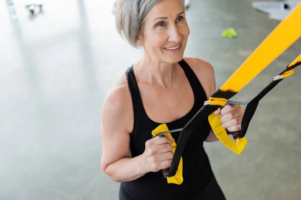 Vue grand angle de femme âgée gaie en tenue de sport noire s'exerçant avec des bretelles de suspension dans la salle de gym — Photo de stock