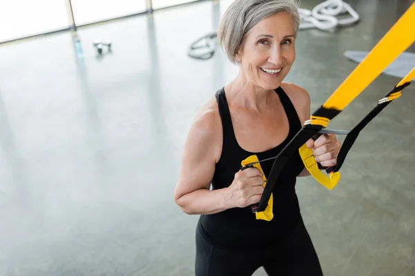 Blick aus der Vogelperspektive auf eine glückliche Seniorin in schwarzer Sportbekleidung, die mit Federbeinen im Fitnessstudio trainiert — Stockfoto