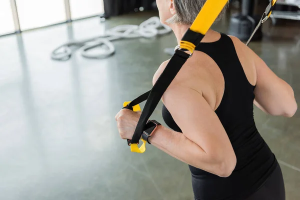 Обрезанный вид пожилой женщины в черной спортивной одежде тренирующейся с подвесными ремнями в тренажерном зале — стоковое фото