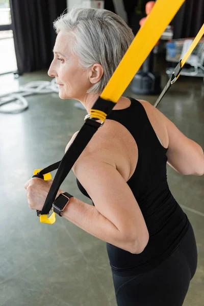 Senior woman in black sportswear exercising with suspension straps in gym — Stock Photo