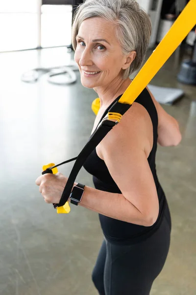 Happy senior woman with grey hair exercising with suspension straps in sports center — Stock Photo