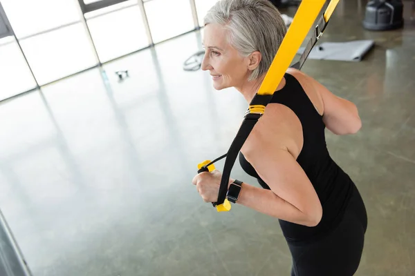 Vue grand angle de la femme âgée souriante en vêtements de sport travailler avec des sangles de suspension dans la salle de gym — Photo de stock