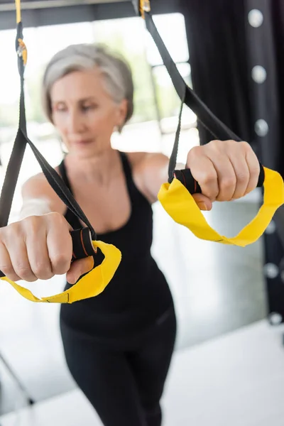 Mulher sênior desfocado em sportswear preto exercício com tiras de suspensão no ginásio — Fotografia de Stock