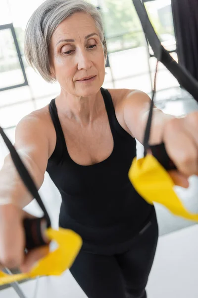 Senior woman with grey hair exercising with suspension straps on blurred foreground — Stock Photo