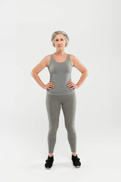 Volle Länge der glücklichen und Rentnerin in Sportbekleidung stehend mit den Händen auf den Hüften auf grau — Stockfoto