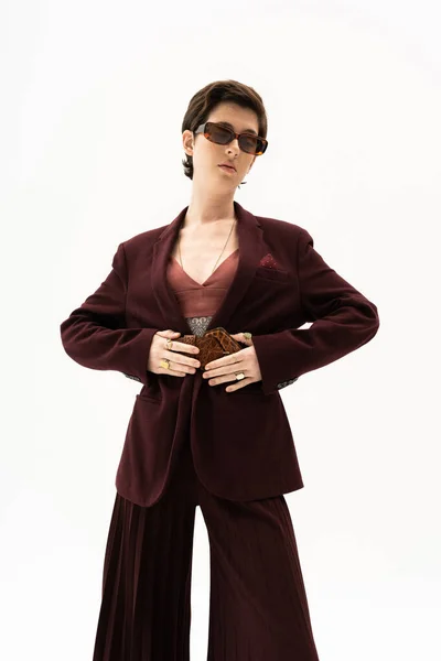Brunette model in burgundy brown pantsuit and sunglasses touching leather belt isolated on white — Stock Photo