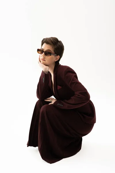 Young model in brown pantsuit and sunglasses posing with hand near face on white background — Stock Photo