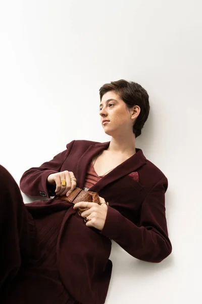 Femme rêveuse en costume à la mode touchant ceinture en cuir et regardant loin sur fond gris — Photo de stock