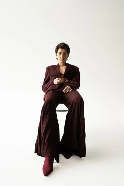 Pleine longueur de femme élégante dans un pantalon large assis sur une chaise et regardant la caméra sur fond gris — Photo de stock