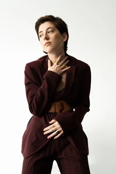 Jovem e sonhadora mulher em blazer e cinto de couro tocando pescoço e olhando para o fundo cinza — Fotografia de Stock