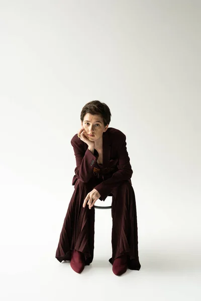 Full length of woman with short brunette hair sitting on chair in elegant pantsuit and looking at camera on grey background — Stock Photo