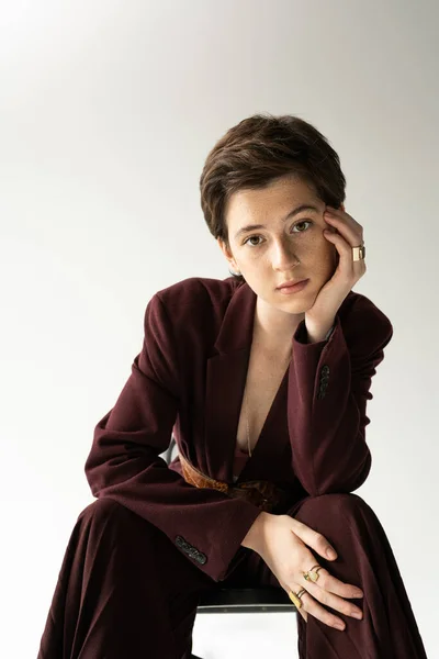 Brunette model in brown suit and finger rings sitting and looking at camera on grey background — Stock Photo