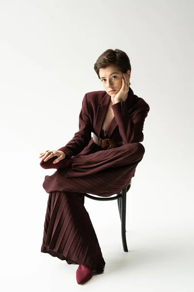 Mujer joven y elegante en pantalones anchos mirando a la cámara mientras posa en silla sobre fondo gris - foto de stock