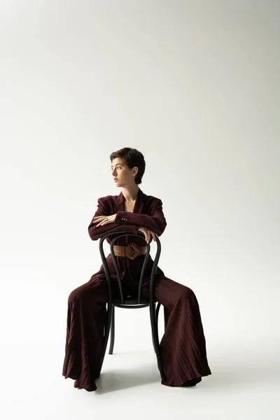 Full length of brunette model in blazer and wide pants sitting on chair and looking away on grey background — Stock Photo