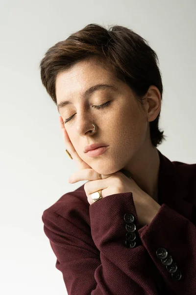 Retrato de una joven pecosa en chaqueta marrón posando con los ojos cerrados aislados en gris - foto de stock