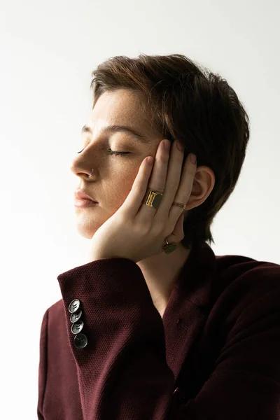 Bonita mujer en blazer marrón burdeos y anillos de dedo posando con los ojos cerrados y la mano cerca de la cara sobre fondo gris - foto de stock