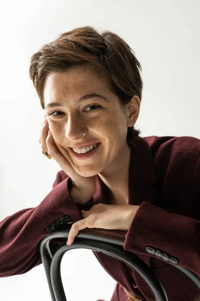 Femme brune insouciante avec des taches de rousseur assis sur la chaise et souriant à la caméra sur fond gris — Photo de stock