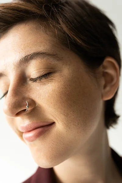 Close up portrait of carefree woman with freckles and nose piercing smiling with closed eyes on grey background — Stock Photo