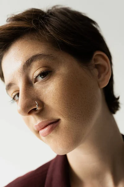 Close up portrait of young freckled woman with nose piercing looking at camera isolated on grey — Stock Photo