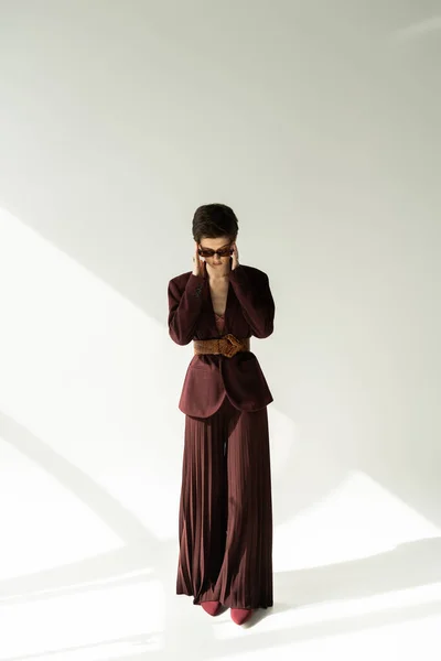 Piena lunghezza del modello bruna in giacca elegante e pantaloni palazzo regolazione occhiali da sole su sfondo grigio con illuminazione — Foto stock