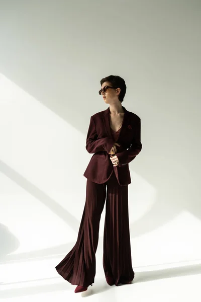 Longitud completa de la mujer morena en gafas de sol y elegante pantsuit fondo gris con iluminación - foto de stock
