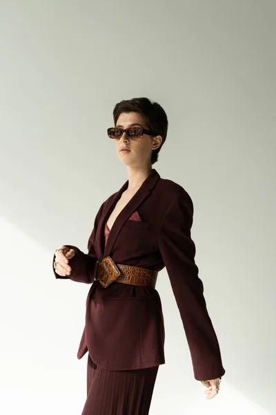 Young brunette woman in sunglasses and brown blazer with leather belt on grey background — Stock Photo