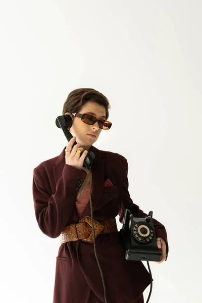 Modello bruna in occhiali da sole e blazer alla moda con cintura in pelle che richiama telefono vintage isolato su grigio — Foto stock