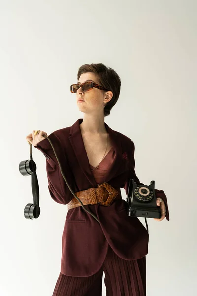 Brunette femme en lunettes de soleil et veste marron bordeaux avec ceinture en cuir posant avec téléphone vintage isolé sur gris — Photo de stock