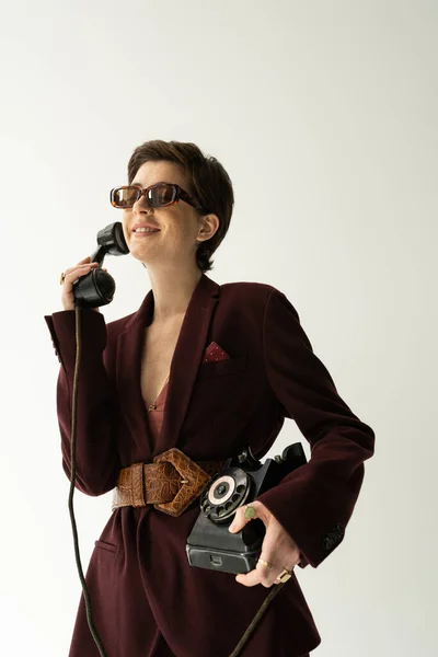 Carefree woman in sunglasses and brown jacket with leather belt talking on retro phone isolated on grey — Stock Photo