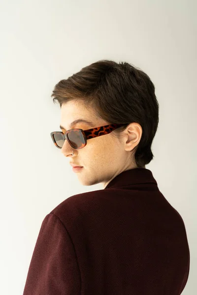 Porträt einer brünetten Frau mit kurzen Haaren, die mit stylischer Sonnenbrille auf grau isoliert posiert — Stockfoto