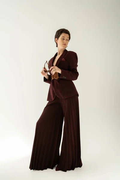 Full length of brunette woman in elegant pantsuit holding sunglasses and looking away on grey background — Stock Photo