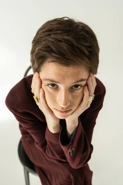 Vista de ángulo alto de la mujer pecosa reflexiva en blazer y anillos de dedos tomados de la mano cerca de la cara y mirando a la cámara en gris - foto de stock