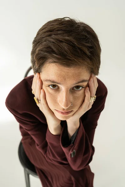 Vue grand angle de femme taches de rousseur réfléchie dans le blazer et les anneaux des doigts tenant les mains près du visage et regardant la caméra sur le gris — Photo de stock