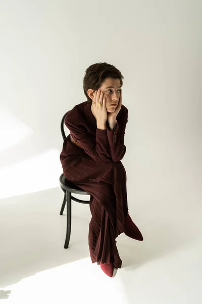 Piena lunghezza di donna alla moda e premurosa in pantaloni larghi seduti sulla sedia e guardando lontano su sfondo grigio con illuminazione — Foto stock