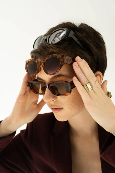 Young woman in finger rings trying on several stylish sunglasses isolated on grey — Stock Photo