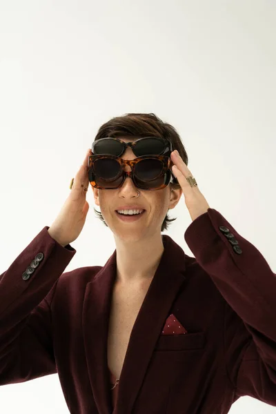 Donna allegra in blazer marrone bordeaux adatta su diversi occhiali da sole alla moda isolati su grigio — Foto stock