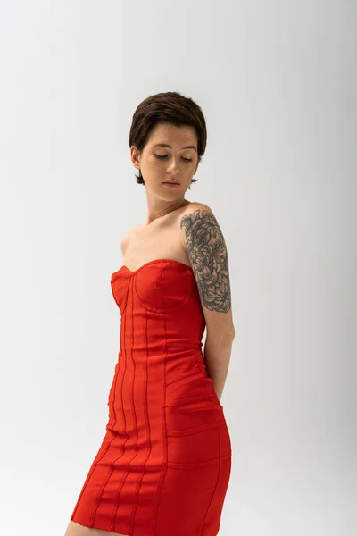 Trendy tattooed woman in red strapless dress posing with hands behind back isolated on grey — Stock Photo