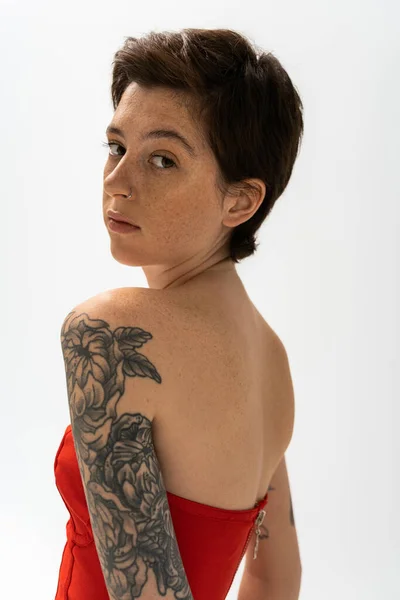 Retrato de mujer joven con tatuaje y pecas mirando a la cámara aislada en gris - foto de stock