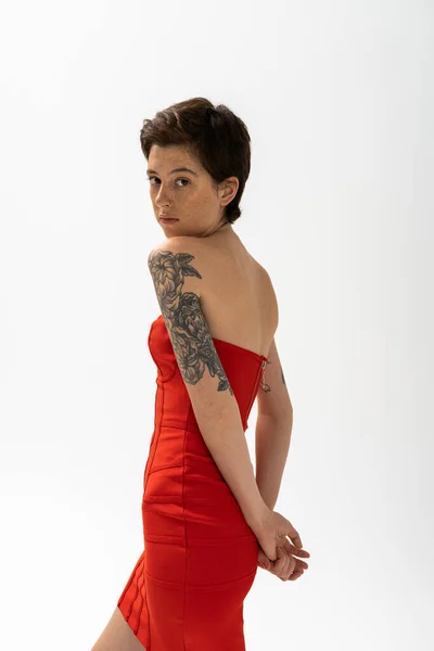 Brunette tattooed woman in red strapless dress standing with hands behind back and looking at camera on grey background — Stock Photo