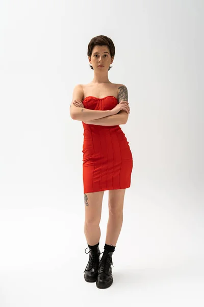 Full length of slender tattooed woman in red strapless dress and black boots posing with crossed arms on grey — Stock Photo