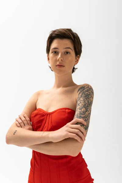 Joven mujer tatuada en vestido rojo sin tirantes cruzando brazos y mirando a la cámara sobre fondo gris - foto de stock