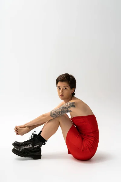 Full length of brunette tattooed woman sitting in red corset dress and rough boots on grey background — Stock Photo